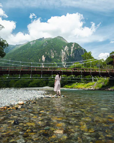 A person in a light dress stands on a rocky riverbank facing a wooden suspension bridge, with tall green mountains and a partly cloudy blue sky in the background.
