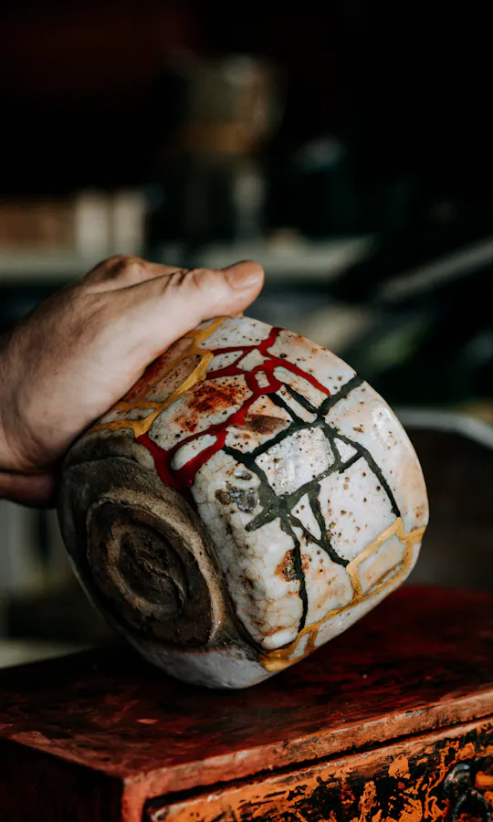 Kintsugi A person's hand holding a large, rustic ceramic bowl with abstract colorful designs, over a weathered red box in a workshop filled with pottery and shelves in the background.