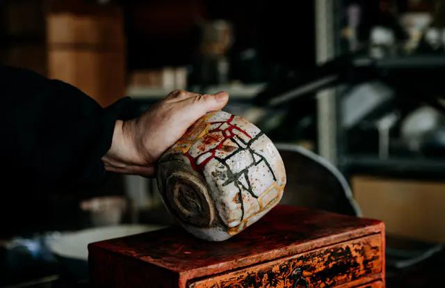 A person's hand holding a large, rustic ceramic bowl with abstract colorful designs, over a weathered red box in a workshop filled with pottery and shelves in the background.