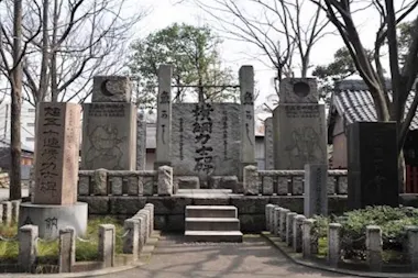 A stone monument with various engraved pillars and statues stands in an outdoor park setting, surrounded by trees and a stone fence, with a small pathway leading up to it. Japanese characters are inscribed on the stones.