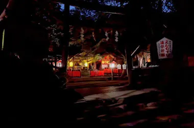 A Japanese shrine illuminated at night, glowing warmly with lanterns and soft yellow lights. The surrounding trees and grounds are dark, creating a tranquil and mysterious atmosphere.