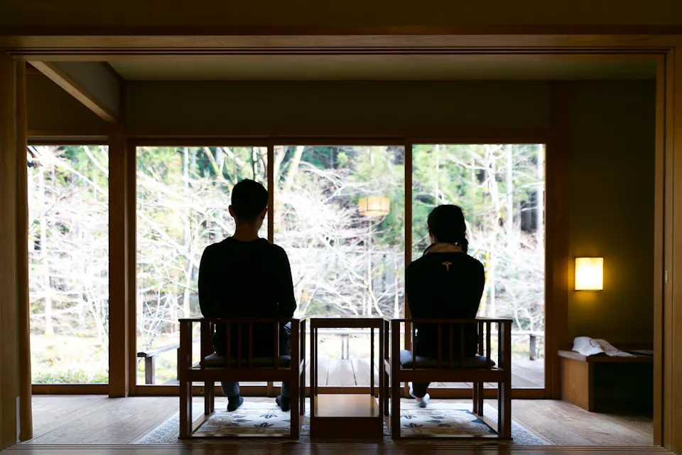 二人は伝統的な日本の部屋で大きな窓を向いて座っています。彼らはクッション付きの木製の椅子に座り、木々と自然の穏やかな景色を眺めています。部屋は暖かい照明と木のアクセントで最小限に装飾されており、穏やかな雰囲気を作り出しています。