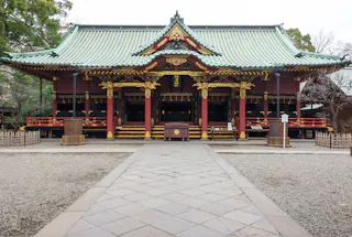A traditional Japanese shrine with ornate gold and red detailing, a green tiled roof, and wooden fencing, viewed from a stone path leading to the entrance. Trees are visible in the background.