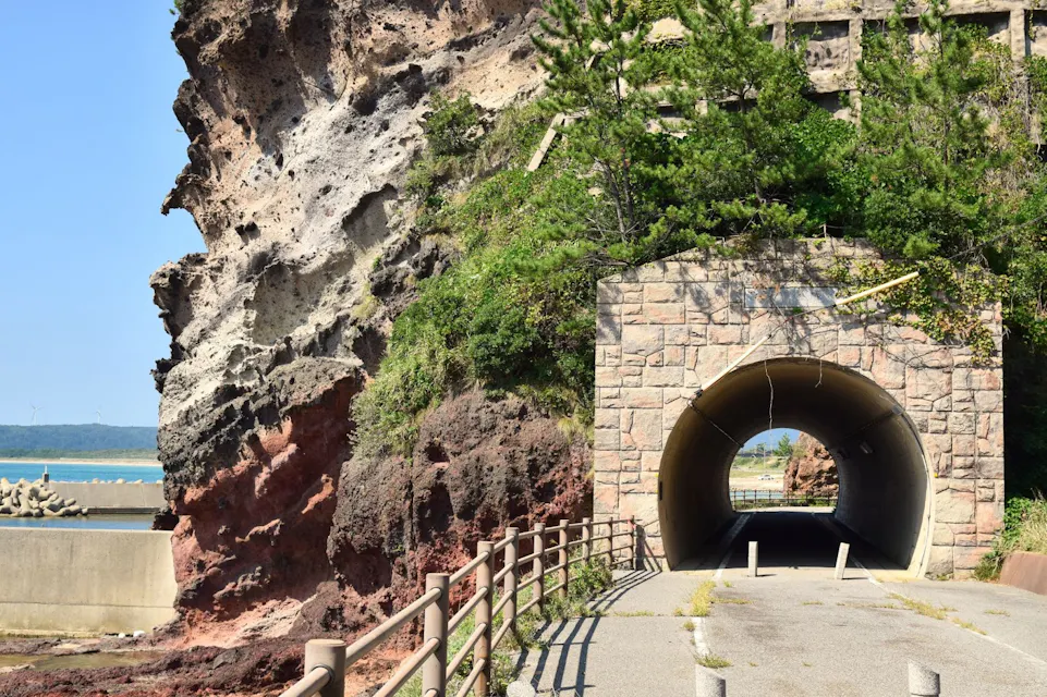 A rocky cliffside with lush greenery stands next to a tunnel made of stone bricks. The entrance to the tunnel is framed by vegetation. A pathway with a metal railing leads to the tunnel, and the ocean is visible in the background.