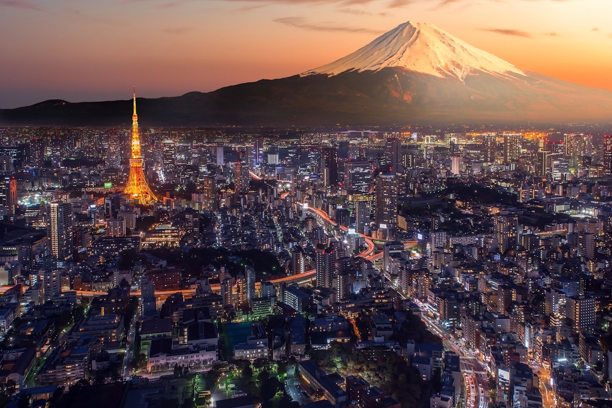 Best Nighttime City Views In Tokyo A vibrant aerial view of Tokyo at dusk showcases the illuminated Tokyo Tower amid a bustling urban landscape. In the background, the iconic Mount Fuji stands majestically with its snow-capped peak glowing under a dramatic sunset sky.