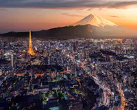A vibrant aerial view of Tokyo at dusk showcases the illuminated Tokyo Tower amid a bustling urban landscape. In the background, the iconic Mount Fuji stands majestically with its snow-capped peak glowing under a dramatic sunset sky.