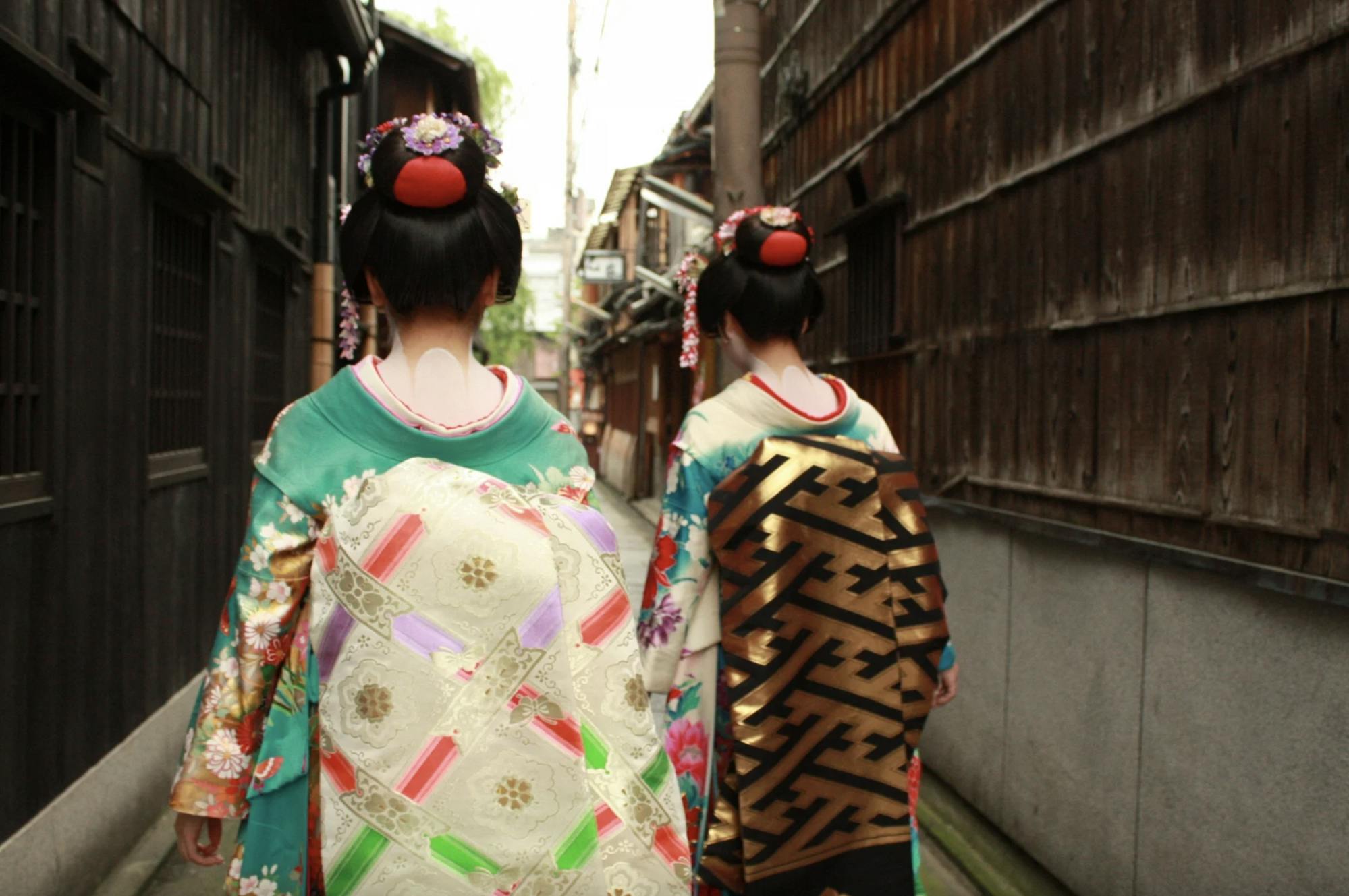 Two people dressed as traditional Japanese maiko in colorful kimonos and elaborate hairpieces walk down a narrow alleyway lined with wooden buildings, seen from behind.