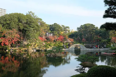 A serene Japanese garden features a curved stone bridge over a reflective pond, surrounded by lush green trees and some autumn foliage. People stroll along the paths, enjoying the tranquil landscape.