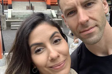 A woman and a man taking a close-up selfie outdoors, smiling softly. There are stone steps, railings, and part of a building structure visible in the background.