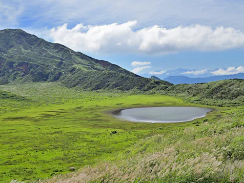 Kusasenrigahama A small, circular lake surrounded by lush green grass and hills under a partly cloudy blue sky, with tall grasses in the foreground and a mountain in the background.