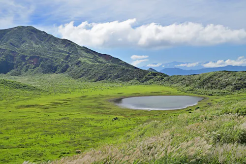 A small, circular lake surrounded by lush green grass and hills under a partly cloudy blue sky, with tall grasses in the foreground and a mountain in the background.