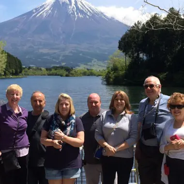 Lake Tanuki A group of seven adults stands smiling by a railing in front of a lake, with trees on both sides and a snow-capped Mount Fuji visible in the background under a partly cloudy sky.