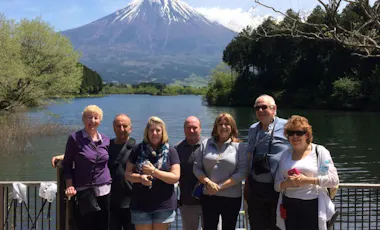 A group of seven adults stands smiling by a railing in front of a lake, with trees on both sides and a snow-capped Mount Fuji visible in the background under a partly cloudy sky.