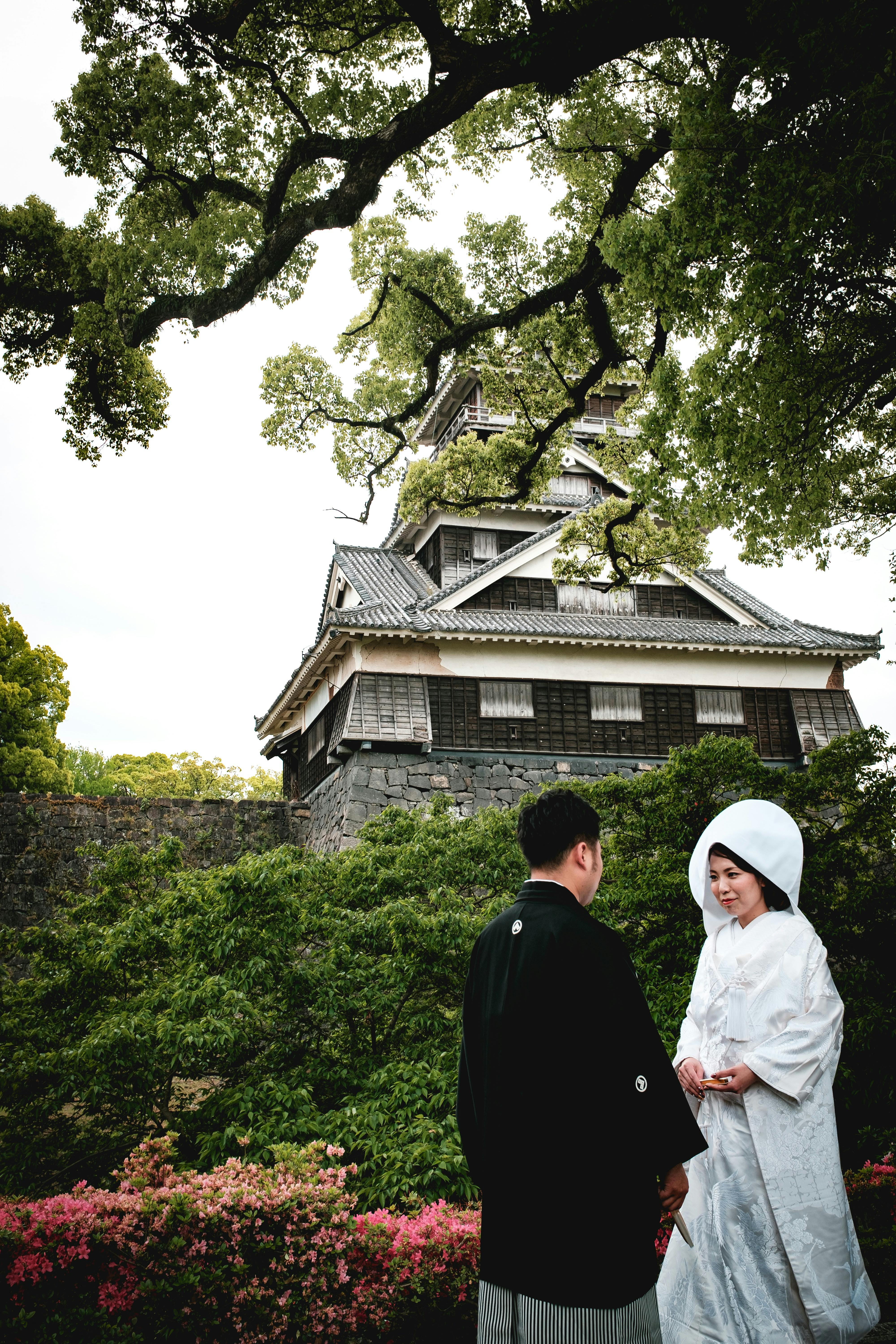 A couple in traditional Japanese wedding attire stands in front of a historic wooden castle, surrounded by lush greenery and colorful flowers. The bride wears a white kimono and headdress, while the groom wears black robes.