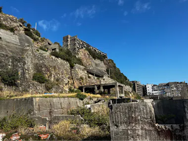Abandoned concrete buildings and overgrown vegetation on a rocky hillside under a clear blue sky, suggesting an old, deserted site.