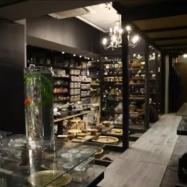 Kintsugi Workshop A cozy, dimly lit store interior with shelves full of pottery and ceramics. In the foreground, tall glass vases with water, green plants, and a goldfish are displayed on clear stands.