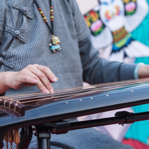 Koto A person wearing a gray outfit plays a traditional string instrument, likely a guzheng or koto. The person's hands gently pluck the strings, and they are adorned with a beaded necklace featuring a small pendant. A colorful backdrop is visible.