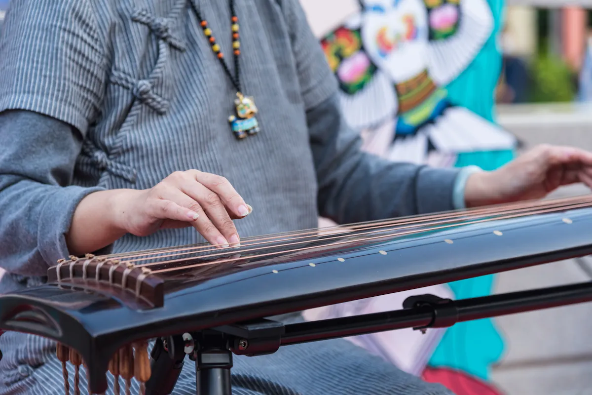 Koto A person wearing a gray outfit plays a traditional string instrument, likely a guzheng or koto. The person's hands gently pluck the strings, and they are adorned with a beaded necklace featuring a small pendant. A colorful backdrop is visible.
