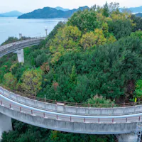 Shimanami Kaido A curving elevated road winds around a lush, green hilltop, overlooking a calm body of water with distant islands and mountains in the background. The road and surrounding nature create a peaceful, scenic vista.