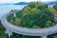 A curving elevated road winds around a lush, green hilltop, overlooking a calm body of water with distant islands and mountains in the background. The road and surrounding nature create a peaceful, scenic vista.