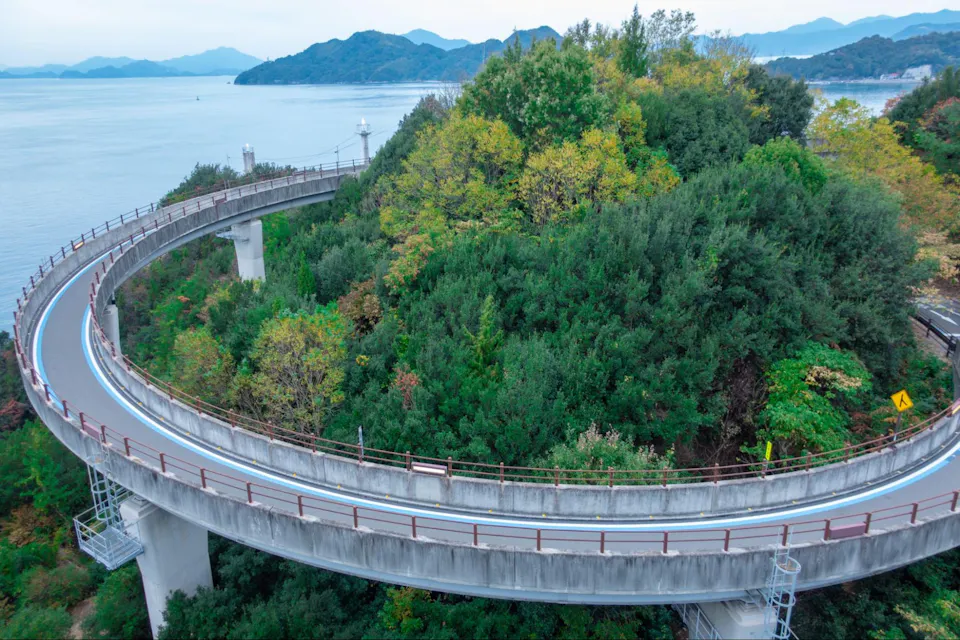 A curving elevated road winds around a lush, green hilltop, overlooking a calm body of water with distant islands and mountains in the background. The road and surrounding nature create a peaceful, scenic vista.