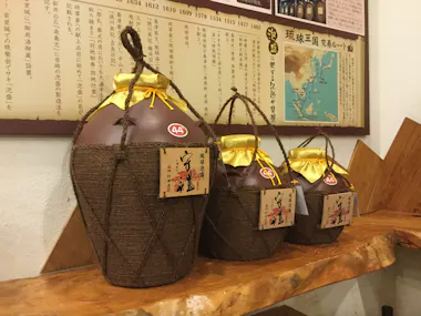 Three large brown ceramic jars wrapped in twine with yellow cloth lids sit on a wooden shelf. Each jar has a tag with Japanese writing. A map and Japanese text are displayed on the wall behind them.