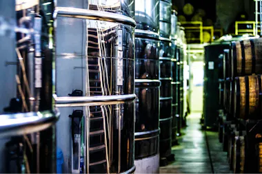 Rows of large metal tanks and wooden barrels line an industrial facility, possibly a brewery or distillery, with pipes and equipment visible in a dimly lit environment.