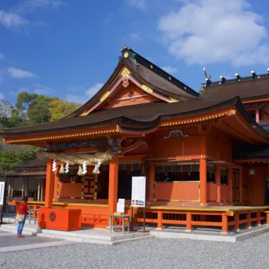Fujisan Hongu Sengen Taisha A traditional Japanese Shinto shrine with bright red-orange wooden architecture, ornate roof decorations, and white paper streamers, set against a blue sky with scattered clouds. A person stands in front of the shrine.