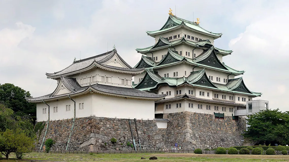 A grand traditional Japanese castle with multiple gabled, green-tiled roofs, sits atop sturdy stone foundations. The building's exterior features white walls and dark wooden accents, surrounded by a green, grassy landscape with a few small trees.