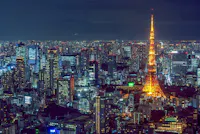 A nighttime aerial view of Tokyo, Japan, featuring a vibrant cityscape illuminated by numerous lights. The brightly lit Tokyo Tower stands prominently in the center, glowing with a warm orange hue against the backdrop of dark buildings and the night sky.