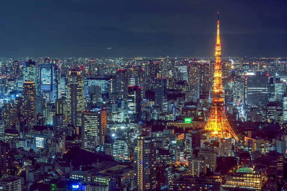 Tokyo Tower at Night A nighttime aerial view of Tokyo, Japan, featuring a vibrant cityscape illuminated by numerous lights. The brightly lit Tokyo Tower stands prominently in the center, glowing with a warm orange hue against the backdrop of dark buildings and the night sky.