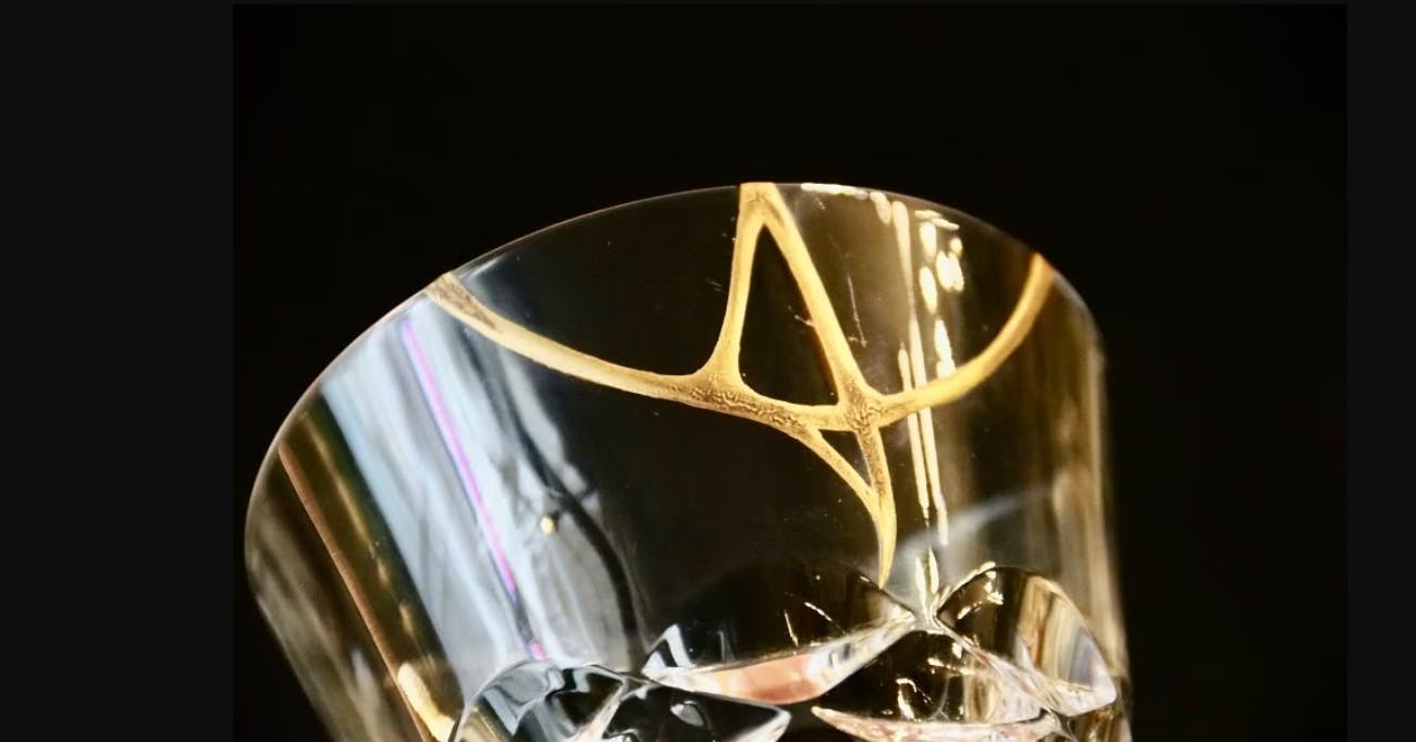 A close-up of a clear glass with a gold decorative pattern near the rim, set against a black background. The glass features cut, faceted details at the base, reflecting light.