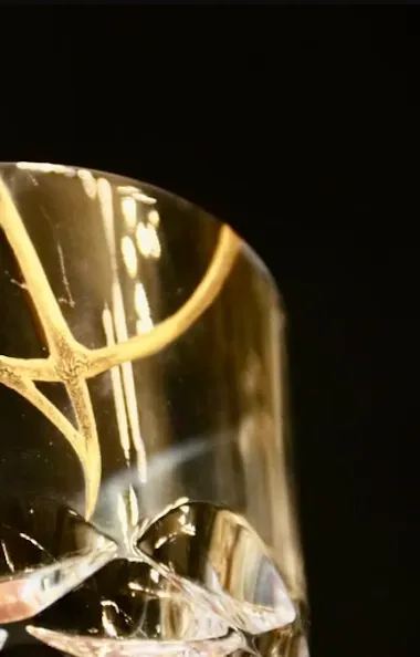 Kintsugi A close-up of a clear glass with a gold decorative pattern near the rim, set against a black background. The glass features cut, faceted details at the base, reflecting light.