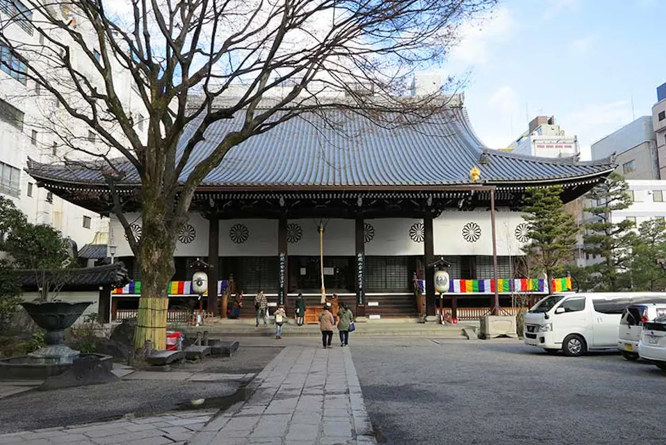 A traditional Japanese temple with a large tiled roof, white walls, and colorful banners near the entrance. Several people are walking in front of the temple. Trees are visible in the courtyard, and buildings surround the temple area. A white van is parked nearby.