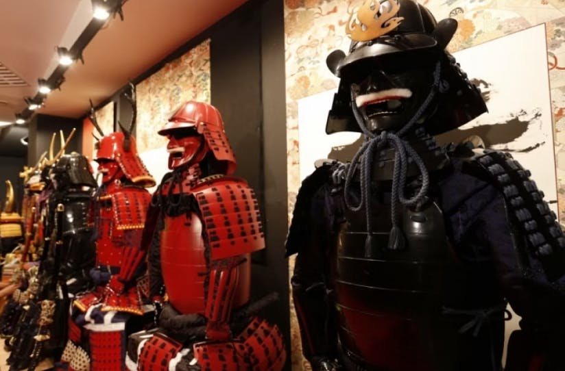 A row of traditional Japanese samurai armor sets, mostly red and black, with helmets and detailed plating, displayed in a museum or exhibition setting with patterned wallpaper in the background.