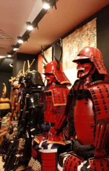 Samurai Armors A row of traditional Japanese samurai armor sets, mostly red and black, with helmets and detailed plating, displayed in a museum or exhibition setting with patterned wallpaper in the background.