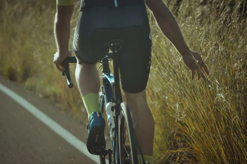 A cyclist wearing a black and green cycling outfit rides a road bike on a rural road. The cyclist's right arm is extended slightly backward, and the left hand is on the handlebars. Grassy vegetation lines the side of the road. The scene is lit by the sunlight.