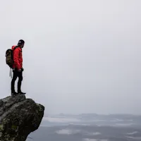 Hiking A person in a red jacket, black hat, and backpack stands on the edge of a cliff overlooking a vast, cloudy landscape. Fog covers the distant mountains, creating a moody and serene atmosphere. The sky is overcast, adding to the dramatic scene.