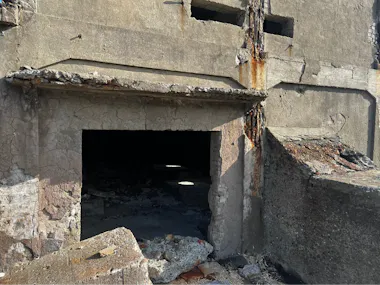 A crumbling concrete building with a large, open doorway leading into darkness; the structure shows signs of severe decay, exposed rebar, cracks, and patches of rust and debris scattered around the entrance.