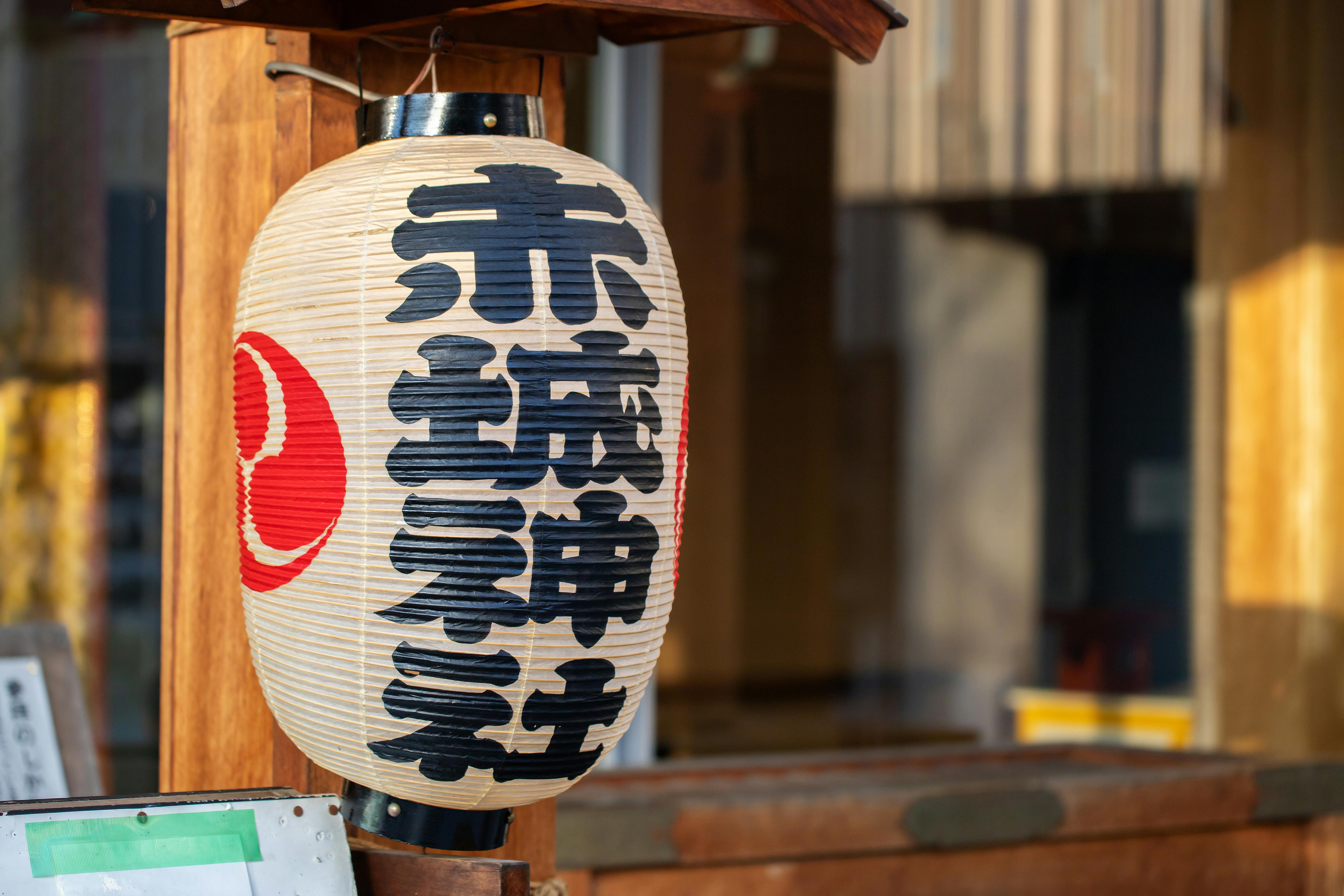 A traditional Japanese paper lantern with bold black kanji characters and a red symbol, hanging outside in sunlight near a wooden structure.