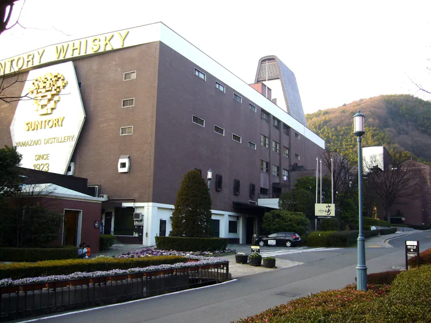 Yamazaki Distillery Image of the Suntory Yamazaki Distillery building, a brown brick structure with the text "Suntory Whisky" on the upper left side. The distillery is surrounded by greenery and hills, with a paved road leading up to the entrance.