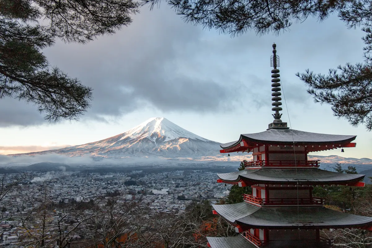 Mount Fuji and Pagoda A scenic view featuring a traditional Japanese pagoda in the foreground and the snow-capped Mount Fuji in the background. The pagoda is surrounded by pine trees, and below is a small town under a cloudy sky.