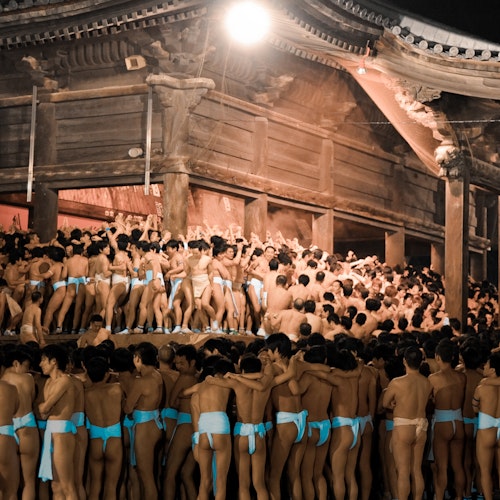 Hadaka Matsuri A large crowd of men wearing traditional white loincloths gather closely together in front of an old wooden temple at night. The scene is brightly lit, highlighting the wooden structure and the dense mass of people beneath the eaves and on the steps.