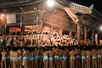 A large crowd of men wearing traditional white loincloths gather closely together in front of an old wooden temple at night. The scene is brightly lit, highlighting the wooden structure and the dense mass of people beneath the eaves and on the steps.