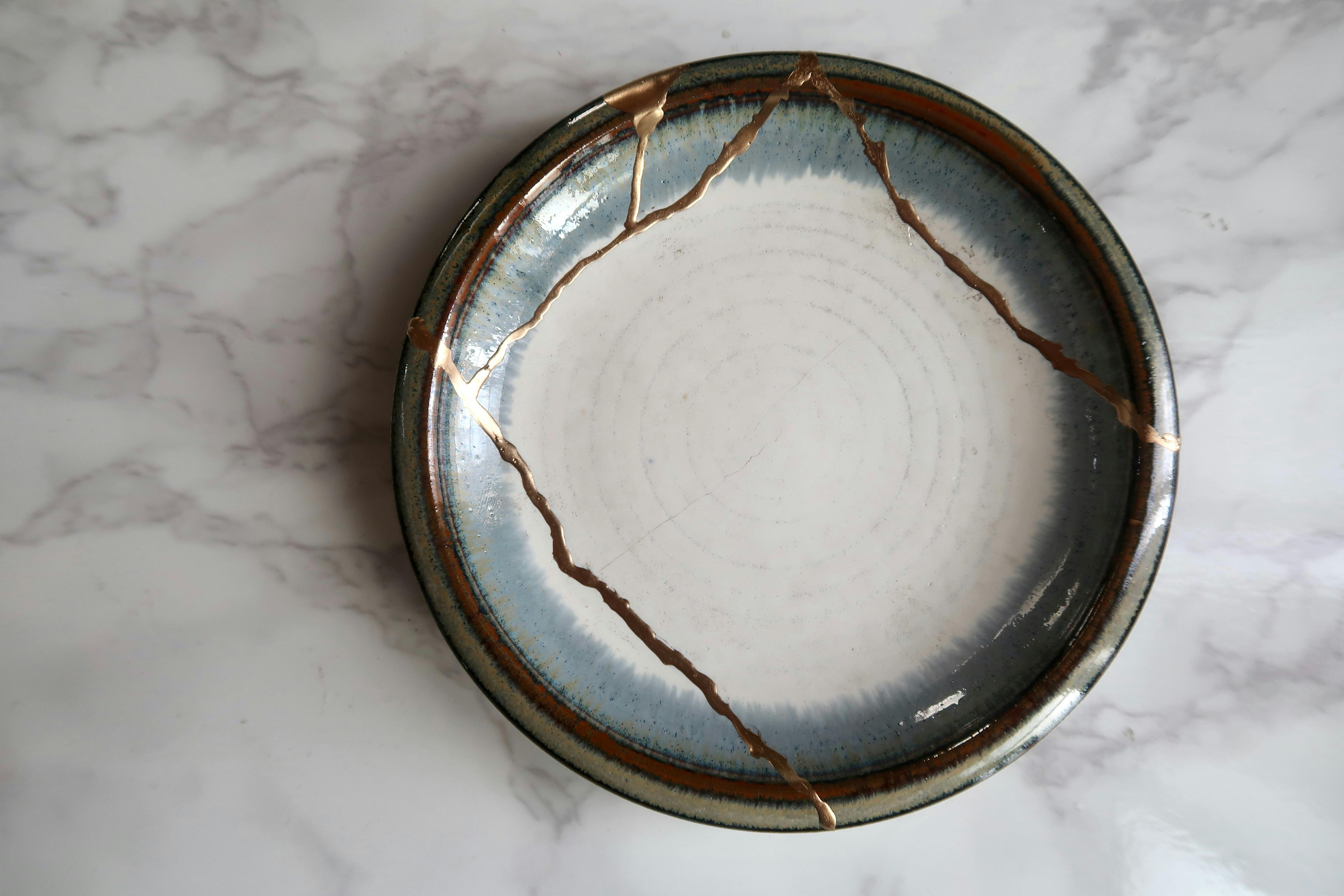 A round ceramic plate with visible cracks repaired using gold, showcasing the Japanese art of kintsugi, placed on a white marble surface.