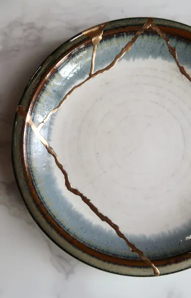 Kintsugi A round ceramic plate with visible cracks repaired using gold, showcasing the Japanese art of kintsugi, placed on a white marble surface.