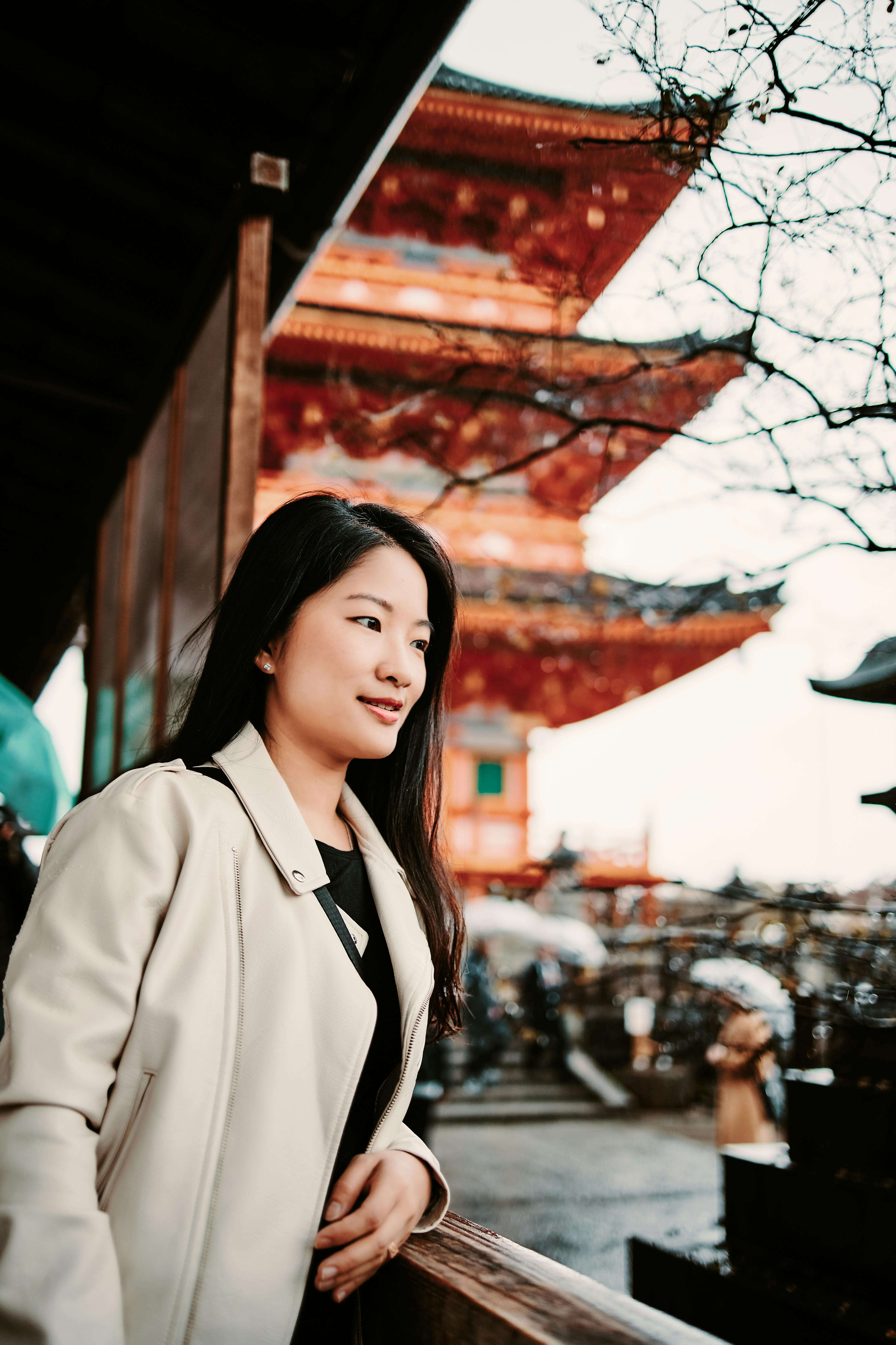 A woman with long dark hair wearing a light-colored coat stands near a wooden railing, with a traditional multi-tiered pagoda and bare tree branches in the background. The scene appears to be at a cultural or historic site.