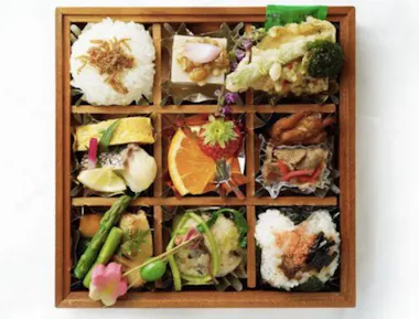 A wooden bento box divided into nine sections, each filled with a variety of Japanese foods including rice balls, tempura, vegetables, fish, fruit, and garnishes, all arranged neatly and colorfully.