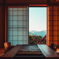 Ryokan A traditional Japanese room with shoji sliding doors that open to a view of mountains. Inside, there's a wooden dining table with chairs on a tatami mat floor. Soft, warm light illuminates the room, creating a tranquil atmosphere.