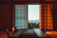A traditional Japanese room with shoji sliding doors that open to a view of mountains. Inside, there's a wooden dining table with chairs on a tatami mat floor. Soft, warm light illuminates the room, creating a tranquil atmosphere.